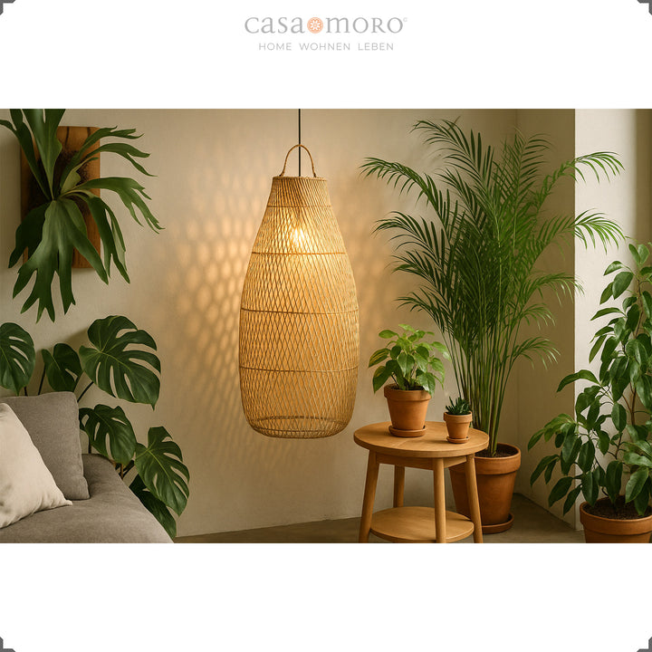 Høj, hængende rattanlampe i stuen med masser af grønne planter og blødt lys